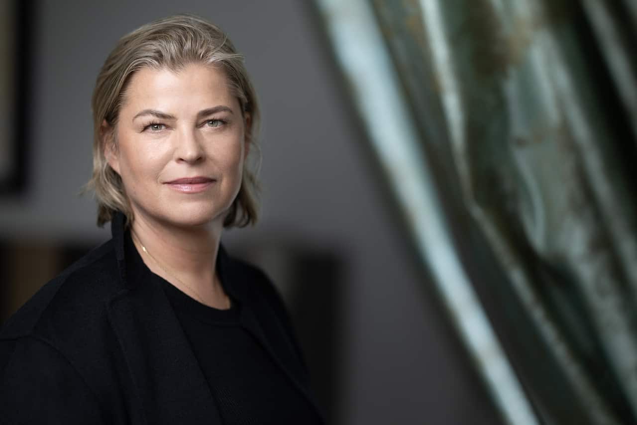 A woman with blonde hair and wearing black clothing stares into the camera 