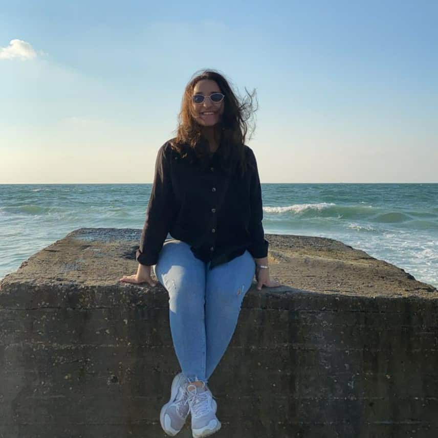 A young woman, dressed in a long-sleeved black shirt, blue jeans, and running shoes, is sitting on a concrete block in front of the sea. She is wearing sunglasses, and her hair blows in the wind.