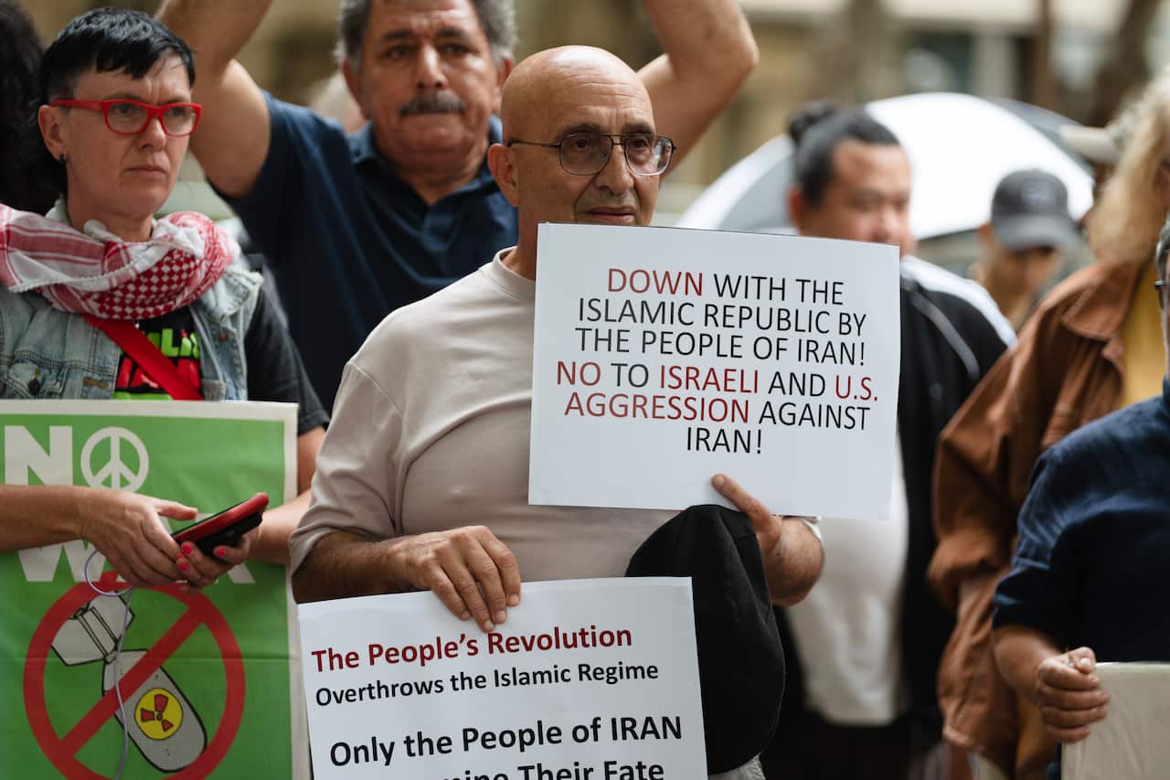 A man at a protest holds signs calling for the people of Iran to overthrow the Islamic Republic while simultaneously opposing military aggression from Israel and the United States.