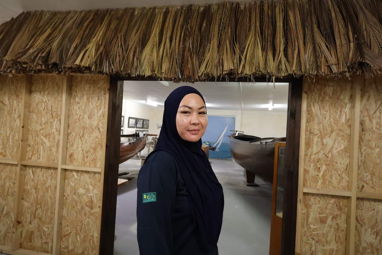 A side-profile of a woman in all-black standing at the entrance of a thatched hut.