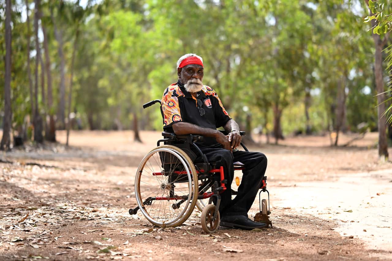 A man in a wheelchair on red dirt. He wears a red headband, black shirt and black pants.