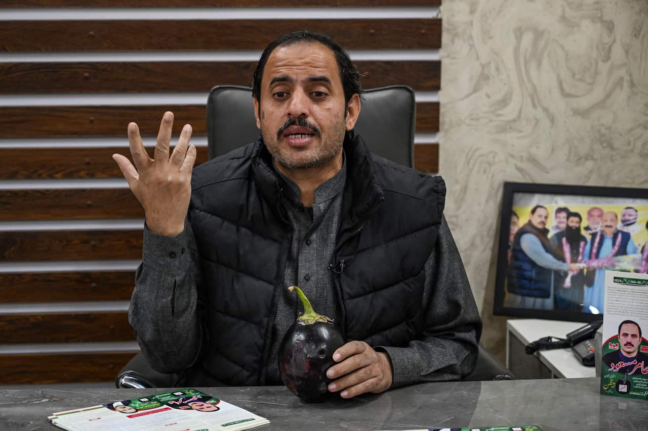 A man in a gray shirt and black vest is sitting at a table with an eggplant in one hand and waving with the other.