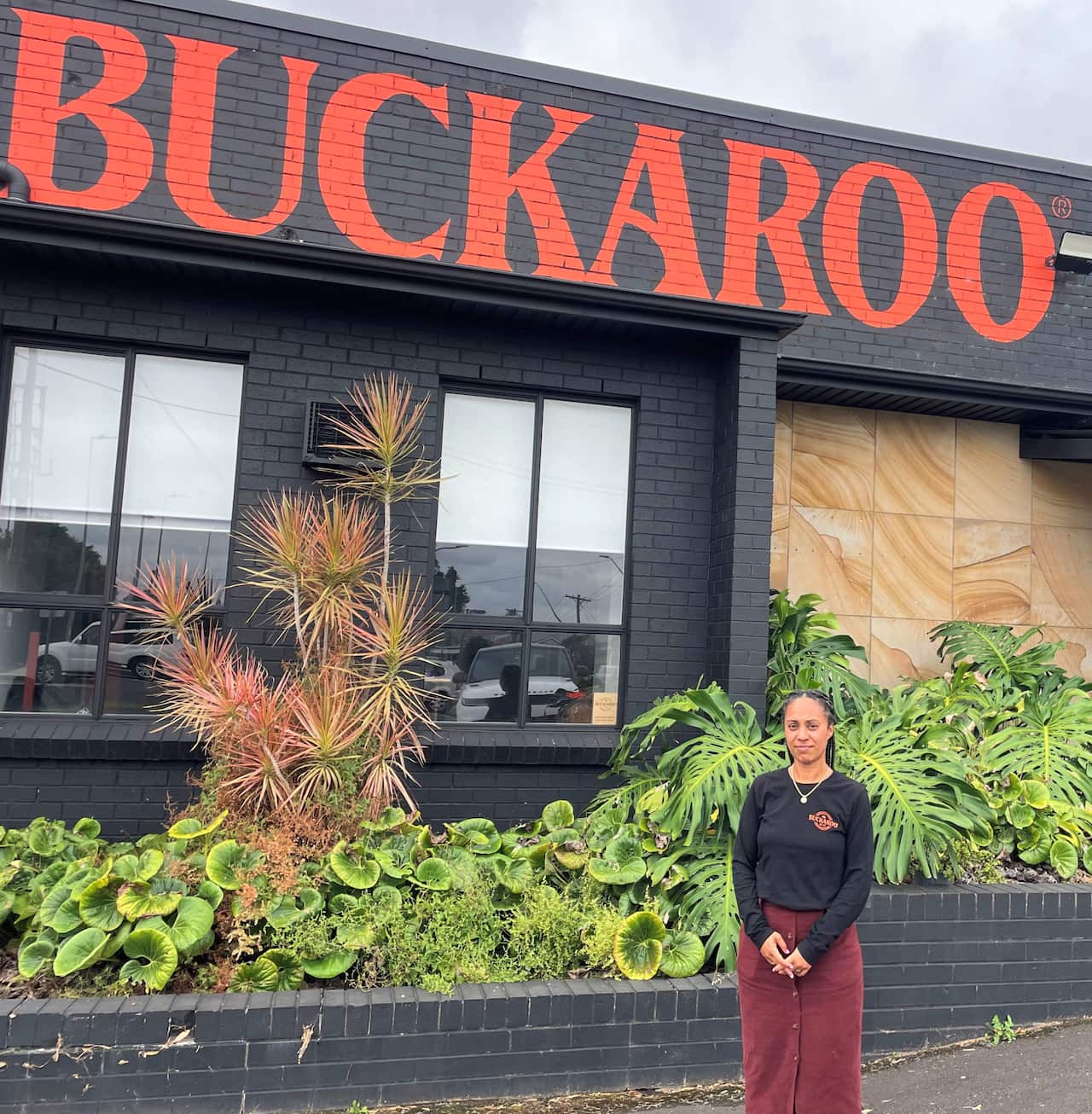 A woman in a black t-shirt and brown skirt stands outside a factory.