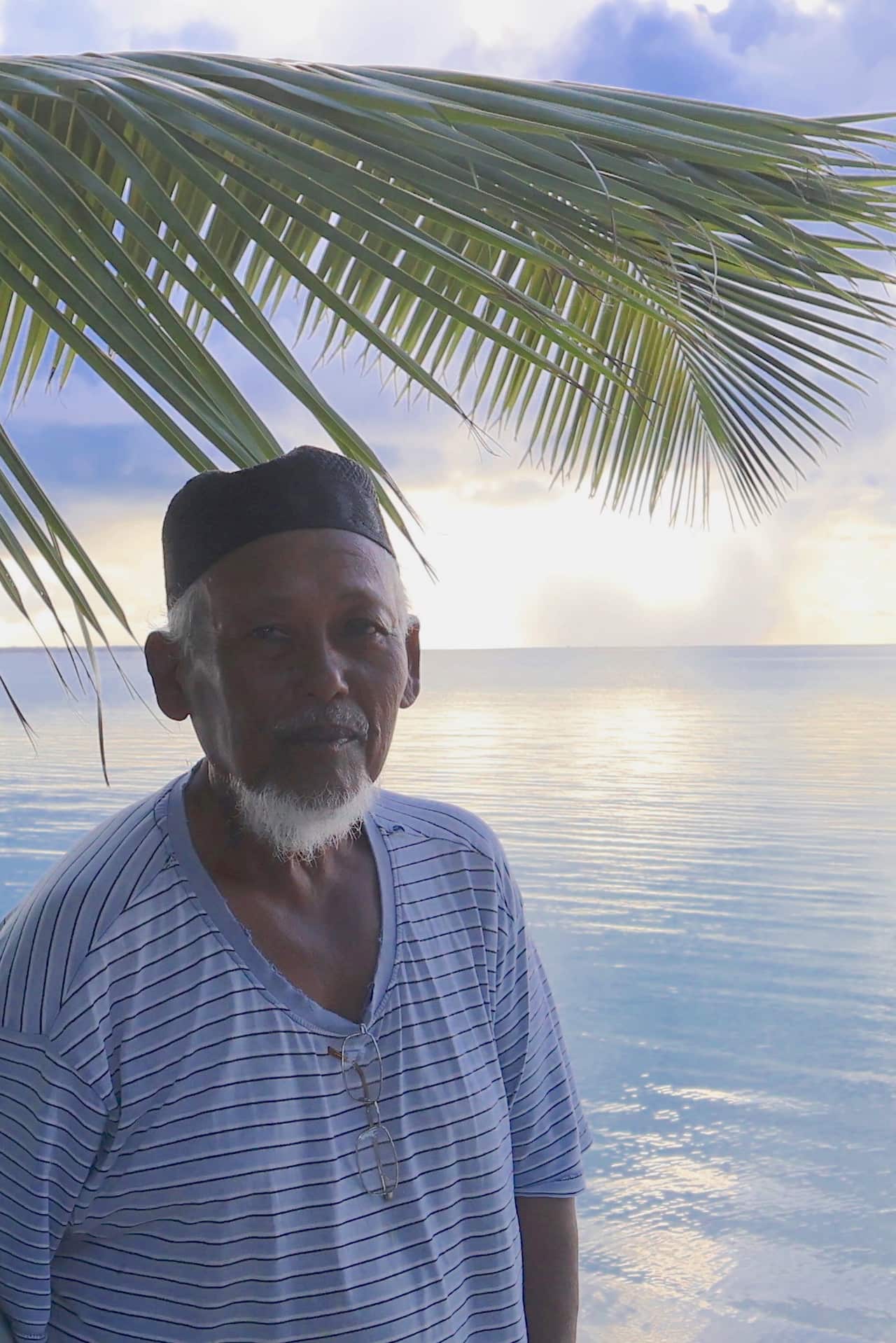 A elderly man stands in front of blue waters with a solemn expression.