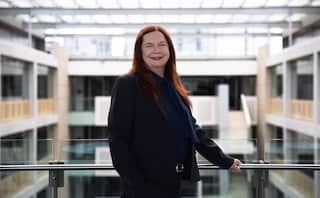 A woman stands near a balcony inside a building. She smiles, while wearing a black suit and has orange hair.