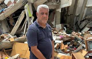 A man with short grey hair and wearing a dark grey polo shirt stands in front of a destroyed building