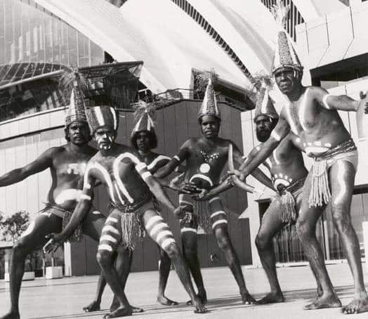 Five Lardil dancers wearing ceremonial dance hats and painted with ochre pose in front of the Opera House. 