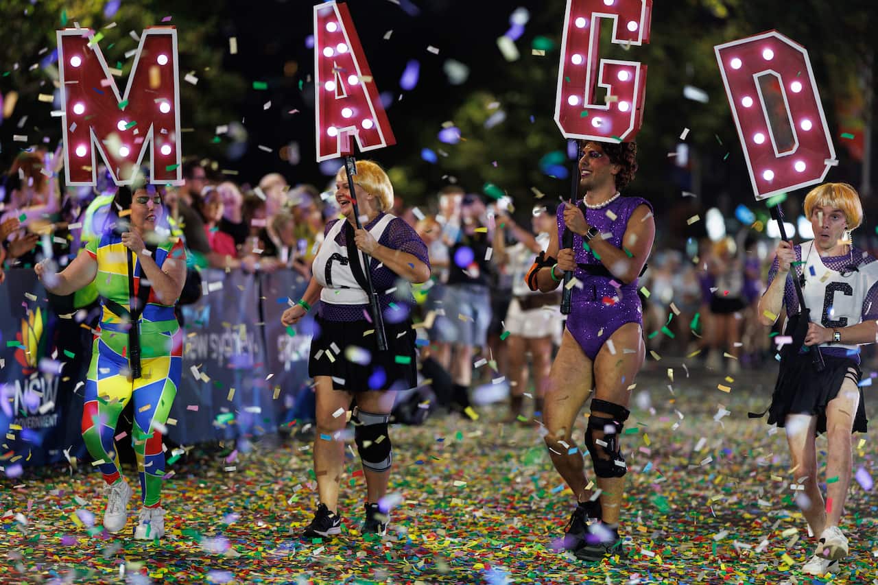 Four performers in colorful costumes march through a cloud of confetti at night. Each holds a large, red illuminated letter, collectively spelling out