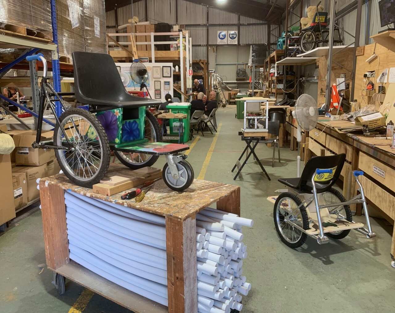 A wheelchair sits on a riser in a workshop with others in the background.