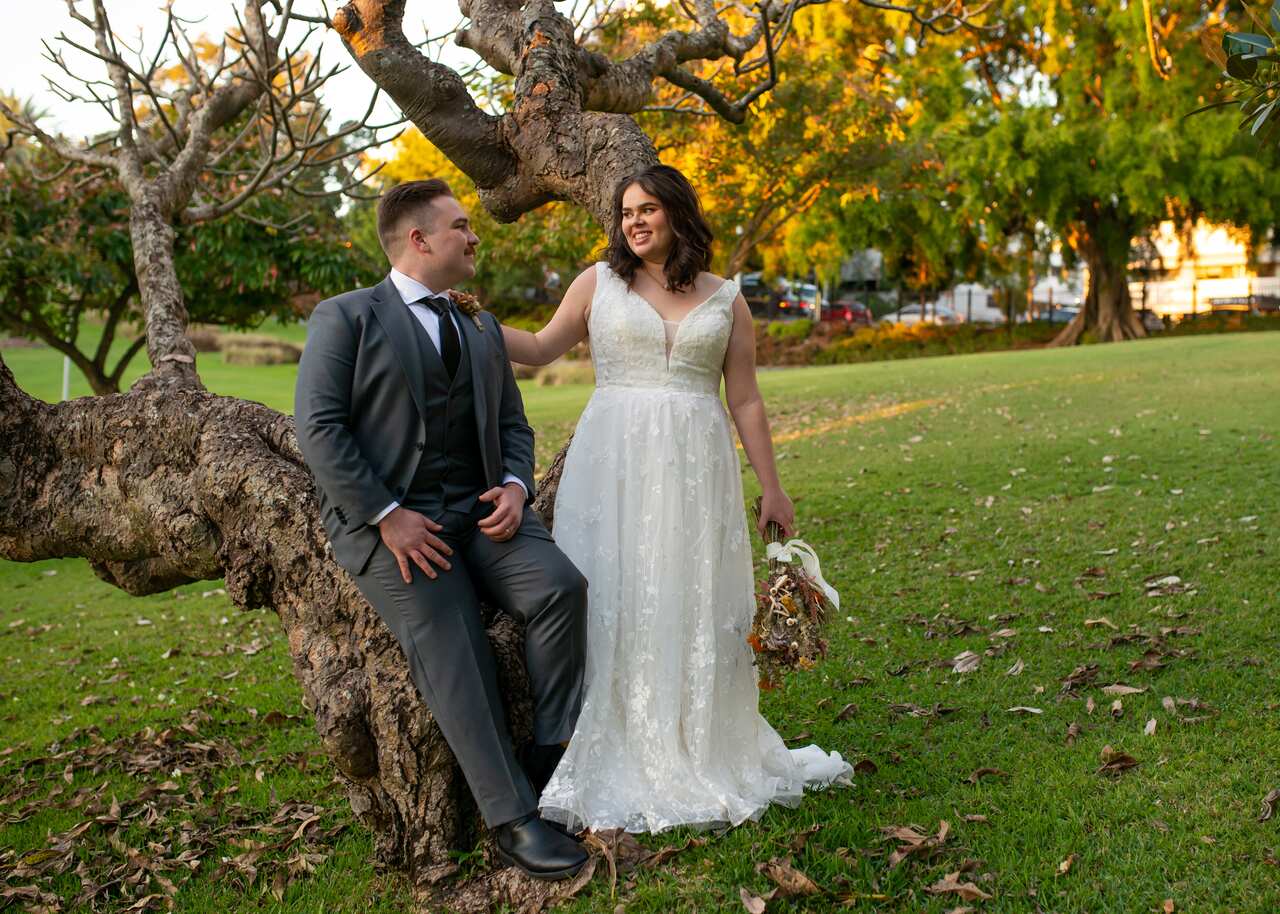 A young couple smile on their wedding day, sitting on a tree in a park.