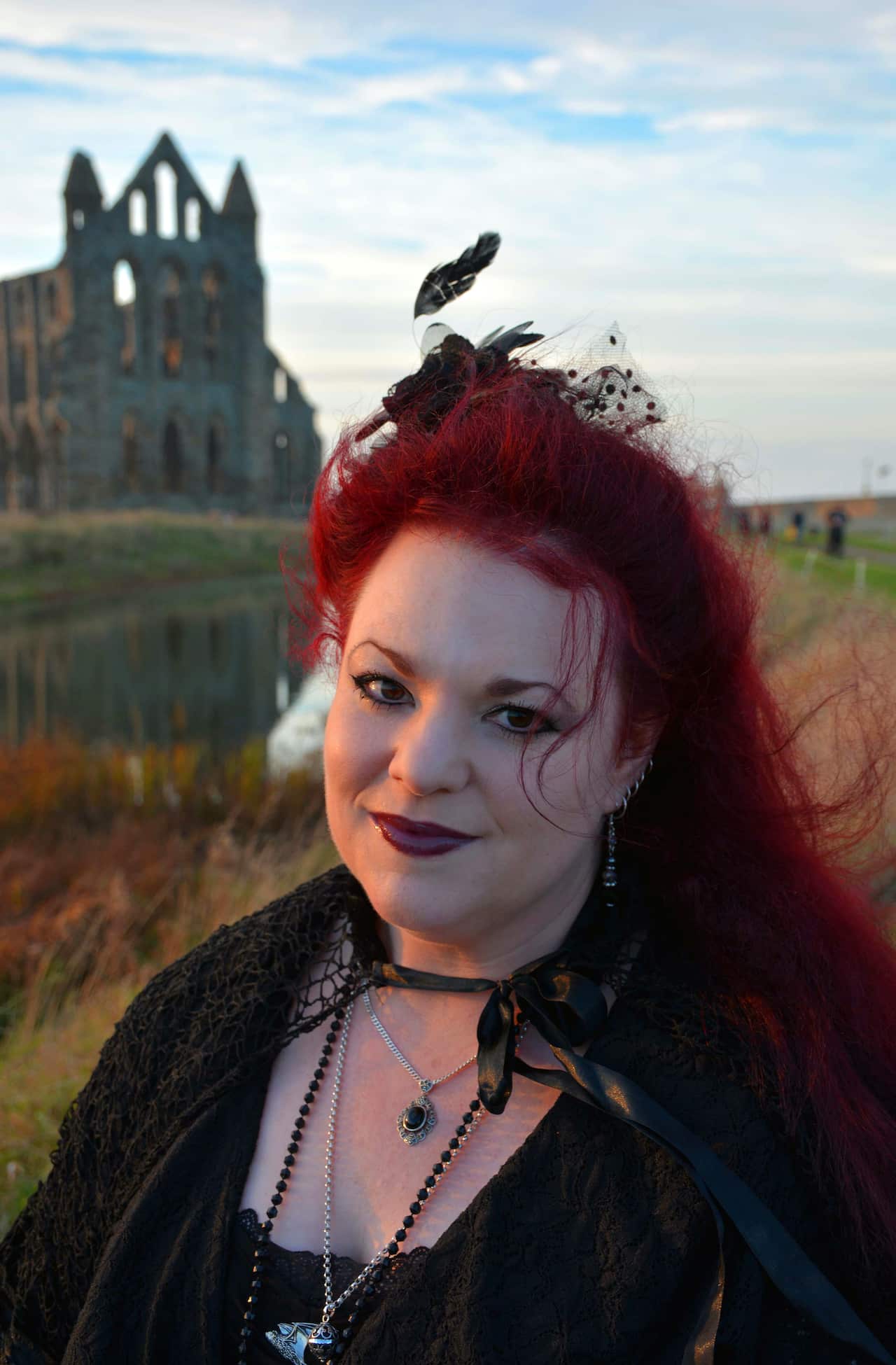 A woman with dyed red hair dressed in black goth clothing stands outside in front of the ruins of a church.