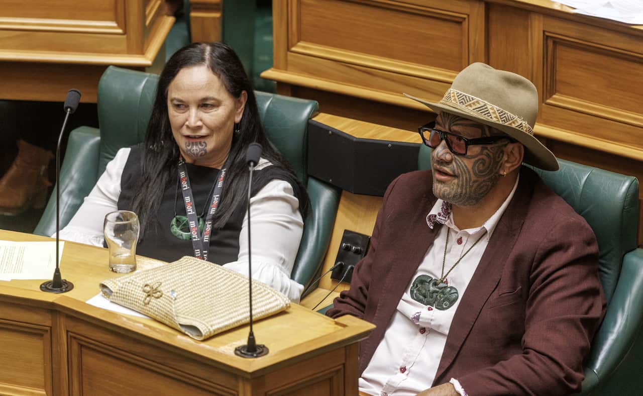 A man and a woman sitting next to each other in green chairs behind a wooden bench inside. The woman has a traditional tattoo on her chin and the man has a traditional tattoo on his face.