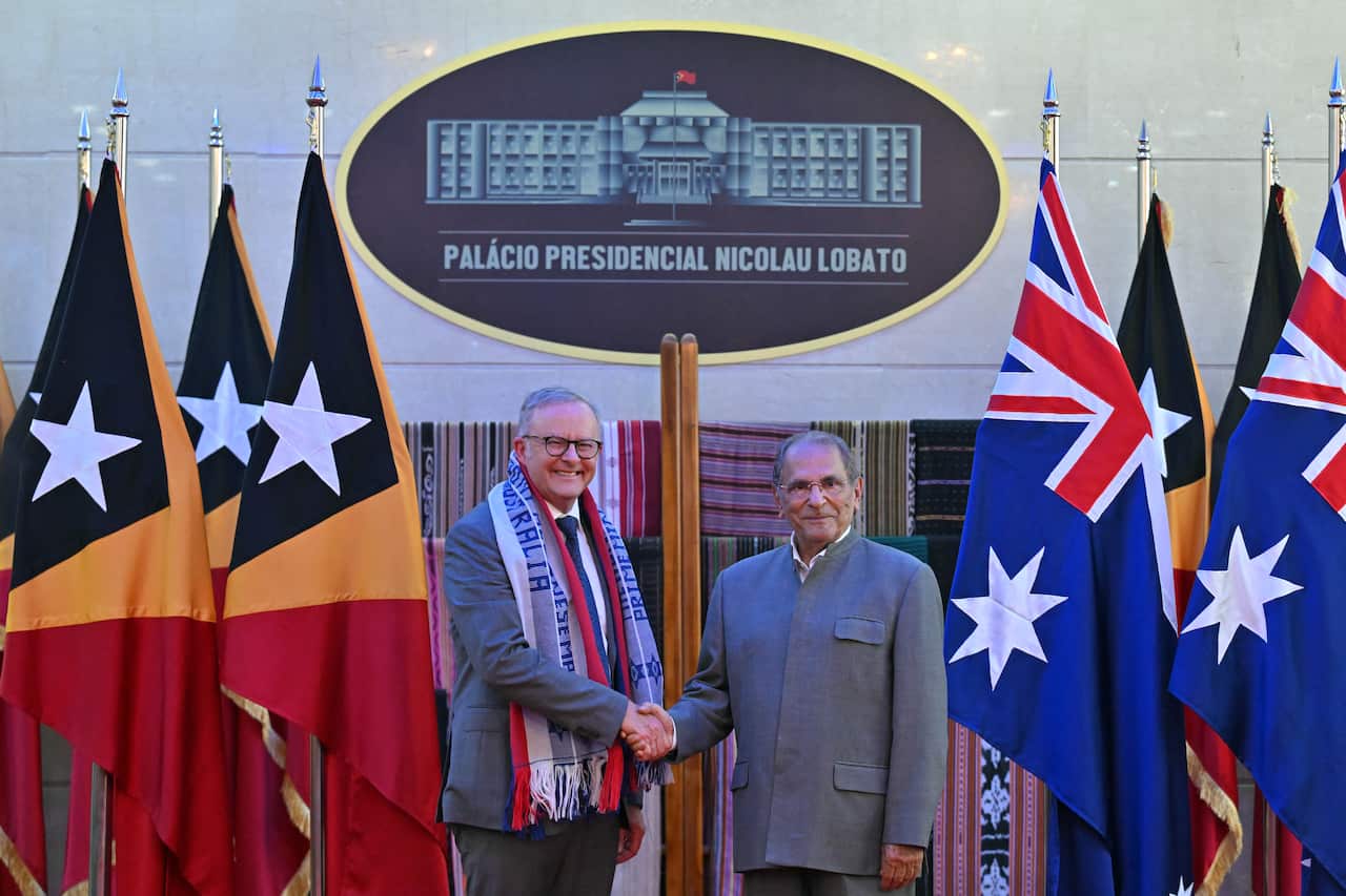 Anthony Albanese and Jose Ramos-Horta shake hands while standing on a podium surrounded by Australian and Timorese flags.