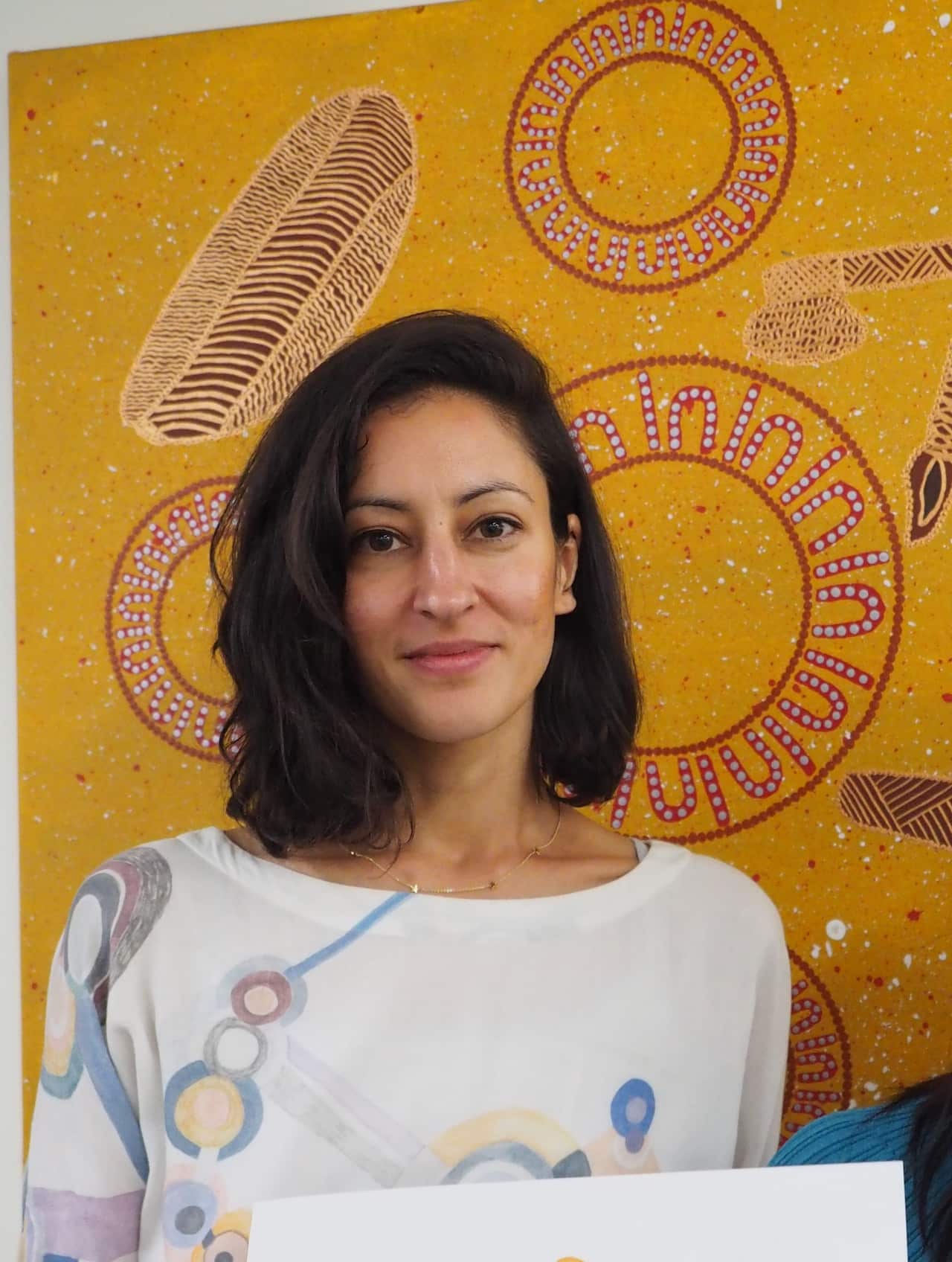 A woman with shoulder-length brown hair wearing a white blouse looks forward with a slight smile.