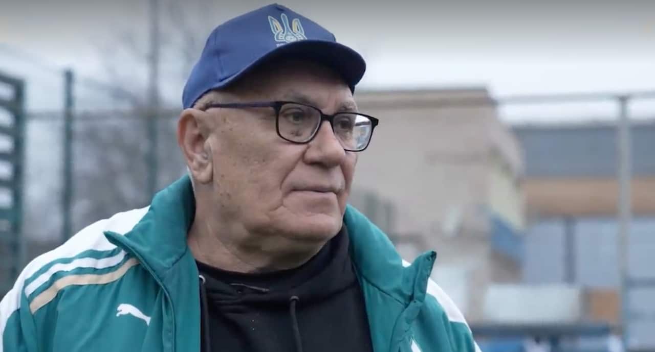 A man wearing a green and white tracksuit top, a blue cap and glasses stands outside with a serious expression on his face.