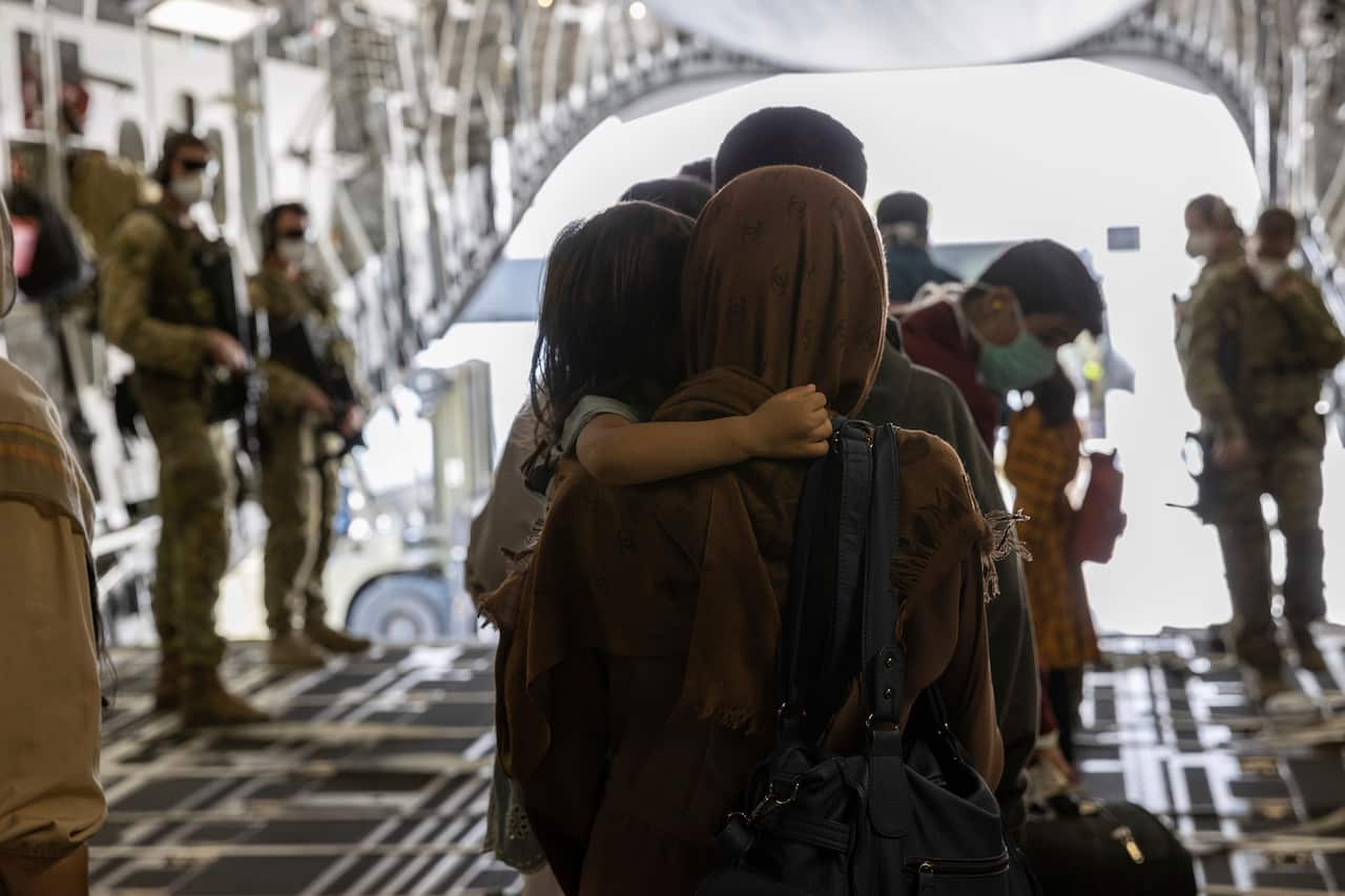 The back of a woman wearing a hijab, with a child's arm wrapped around her. They are standing in a military cargo plane.