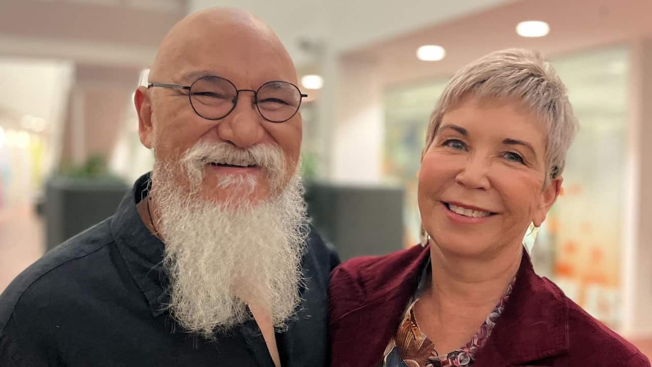 a closeup photo of a middle aged man with a long white beard and glasses and a woman with short blonde hair. The man is wearing a black shirt and the woman is wearing a maroon jacket
