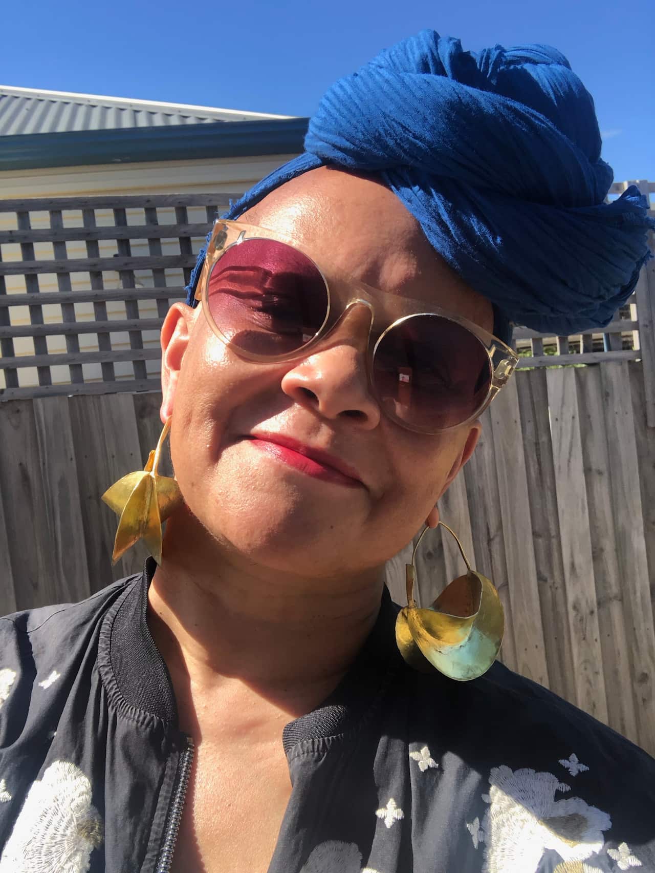 A woman in a blue headwrap, gold earrings, a black and white patterned jacket, sunglasses, and red lipstick.