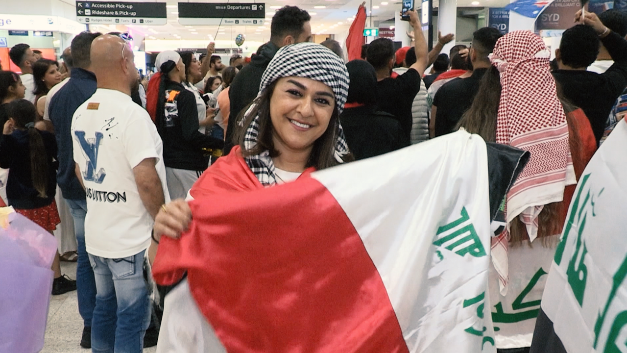 A smiling woman wearing a keffiyeh and an Iraqi flag draped over her shoulders stands among a crowd of people at an airport.