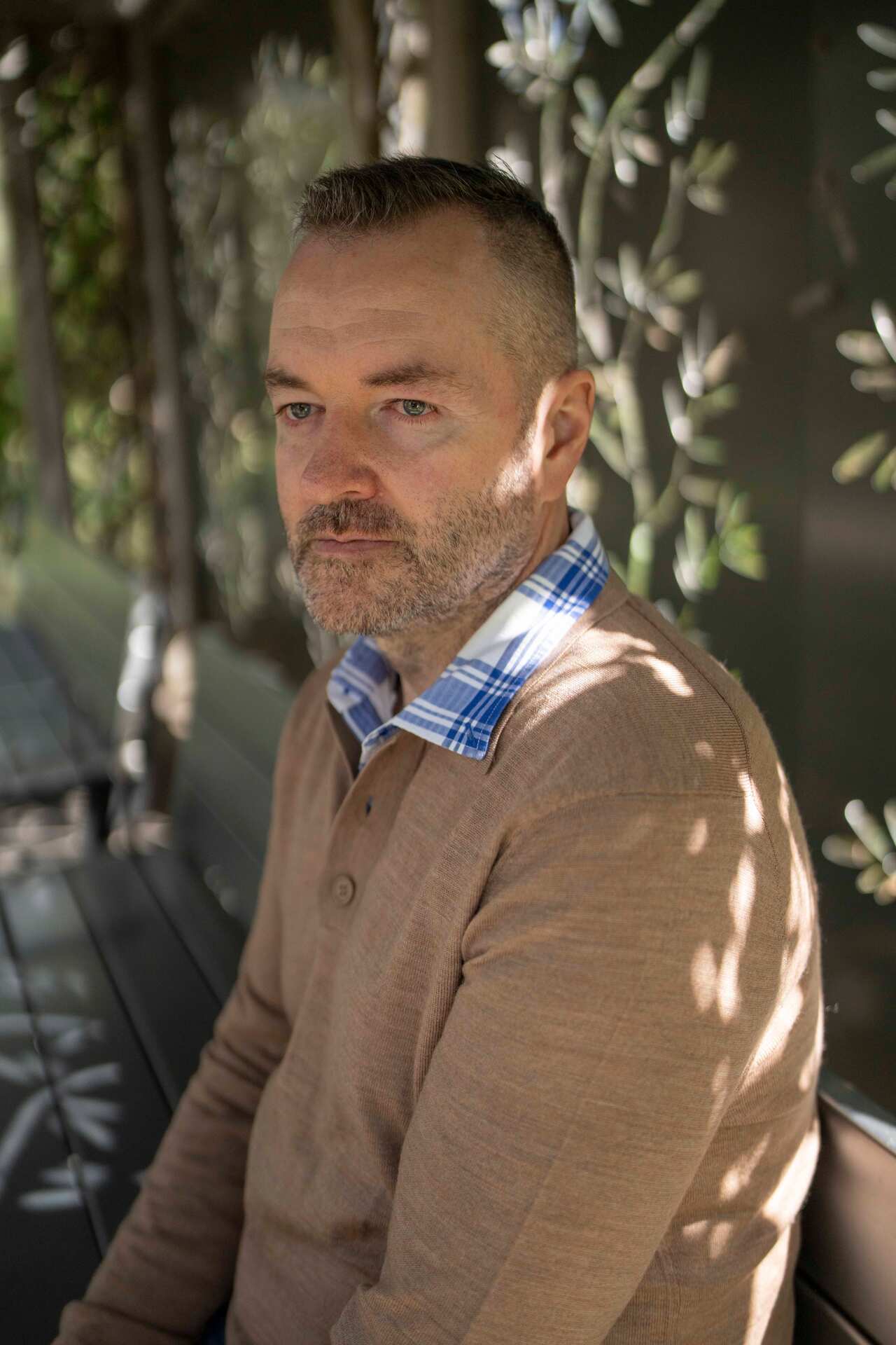 A middle-aged man with greying hair wears a taupe sweater and sits on a bench in a park under dappled light.