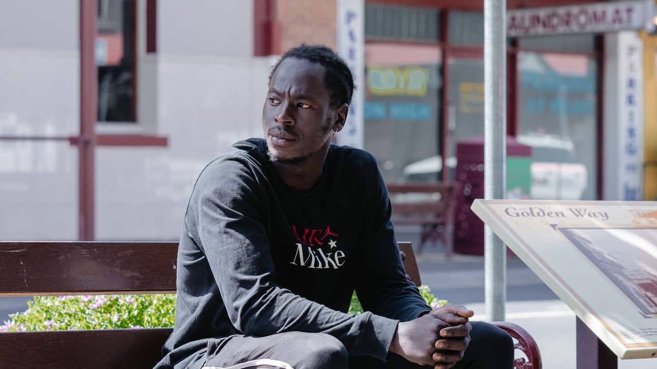 A young South Sudanese man sits on a bench, what looks like a country store can be seen, out of focus, behind him. 