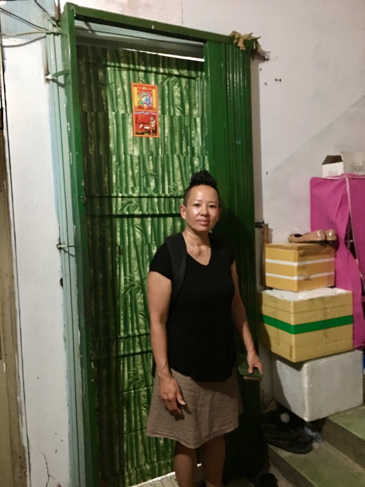 A woman in a black shirt and khaki skirt stands in front of a green door with a pile of boxes next to it. 