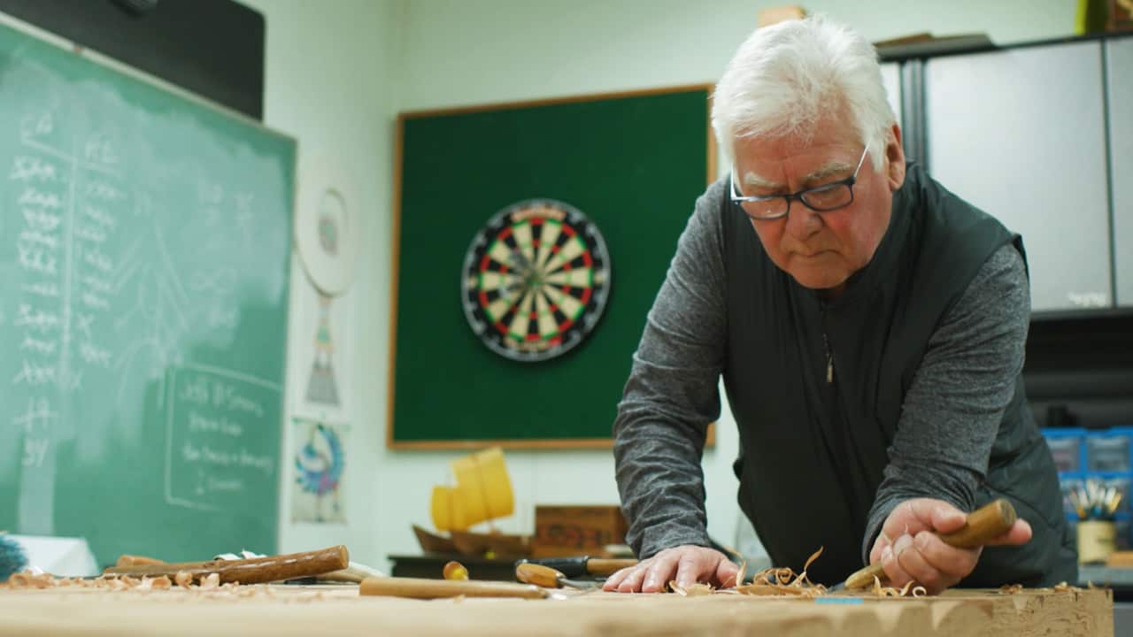 An older man with white hair and glasses and wearing a grey vest is carving wood with a tool in his left hand
