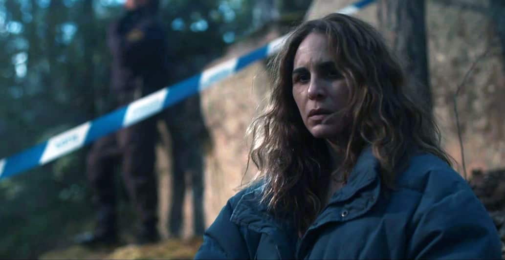 A woman with a worried expression and long loose hair, wearing a blue jacket, stands outdoors. A line of blue and white crime scene tape runs behind her, on an angle.  