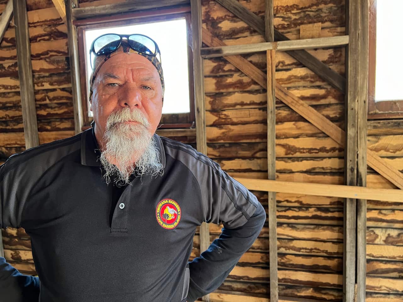 A man with a long grey goatee, sunglasses on his head and wearing a long-sleeved dark grey top stands inside a wooden building