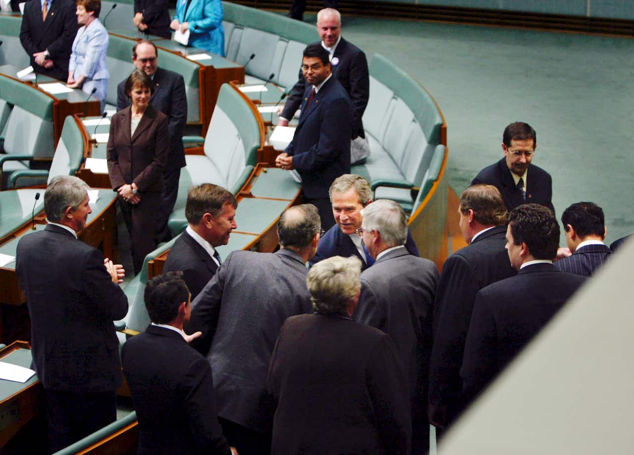 Two men shake hands surrounded by a group of men in suits inside the House of Representatives.