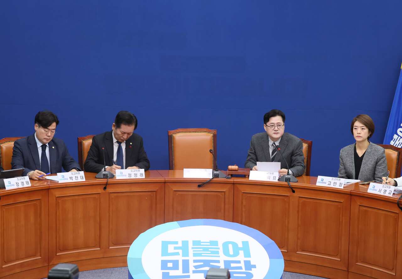 Four people, three men and one woman, dressed formally, sit at a table with an empty chair in the middle. 