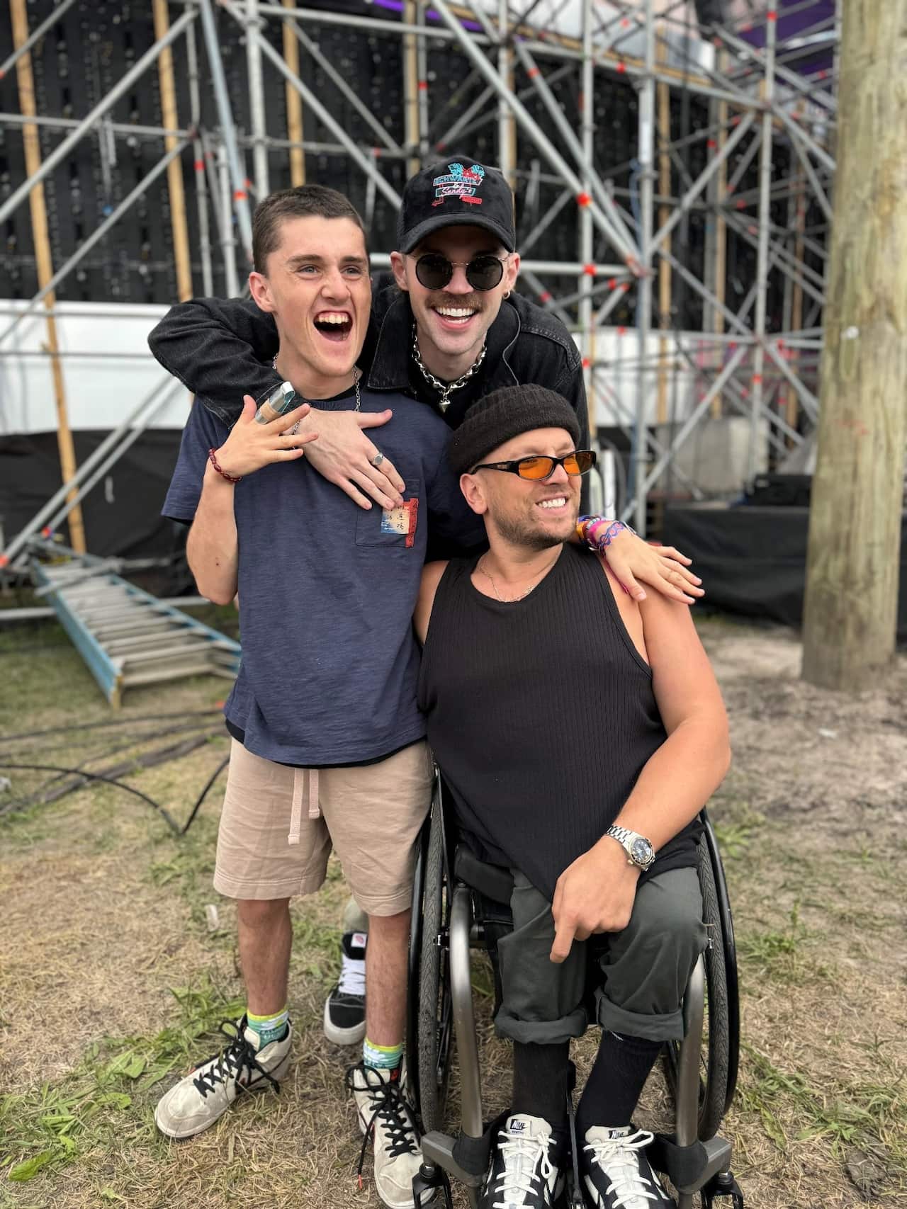 Three men backstage at a music festival. Two are standing with their arms around each other while the other is sitting in a wheelchair