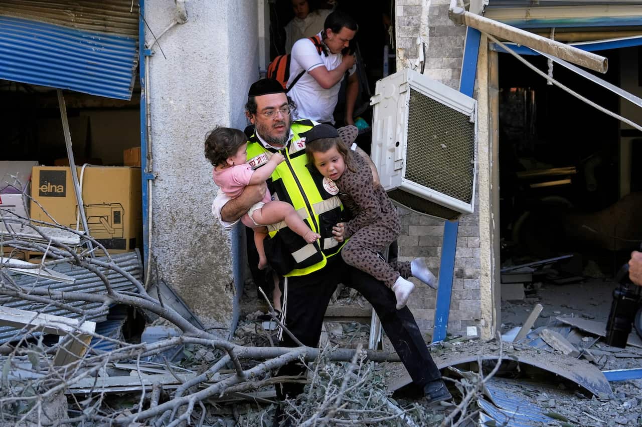 A rescue worker in a hi-vis vest carries two children in his arms out of a damaged building.