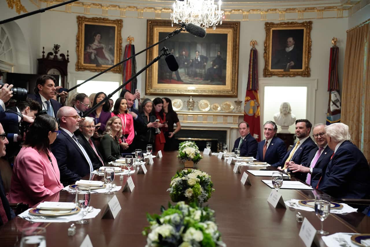 People in business suits sit on either side of a long wooden table with journalists standing on the left.