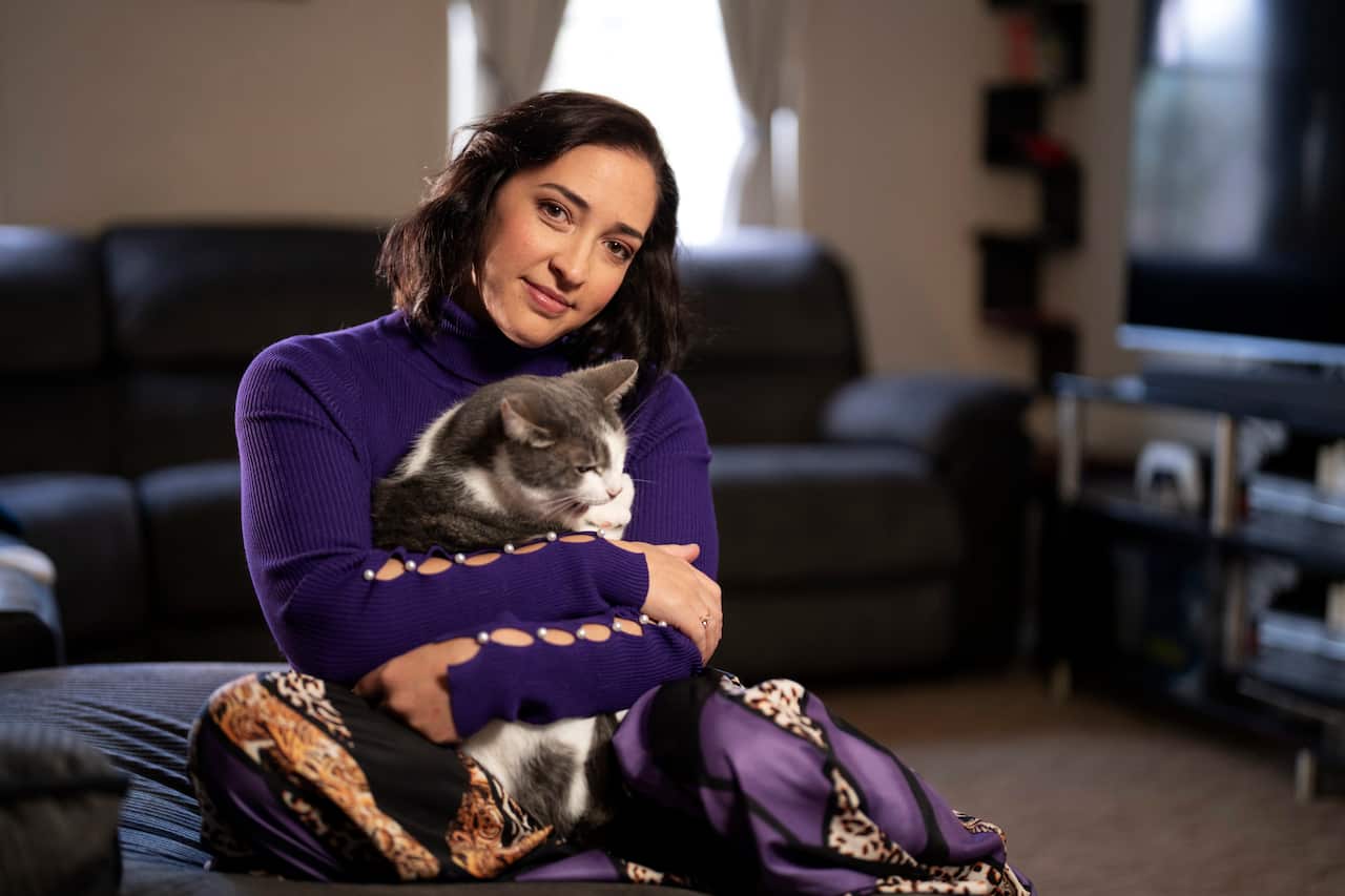 A 30-something white woman with dark brown hair sits at a dining table, wearing a long-sleeved pink shirt.