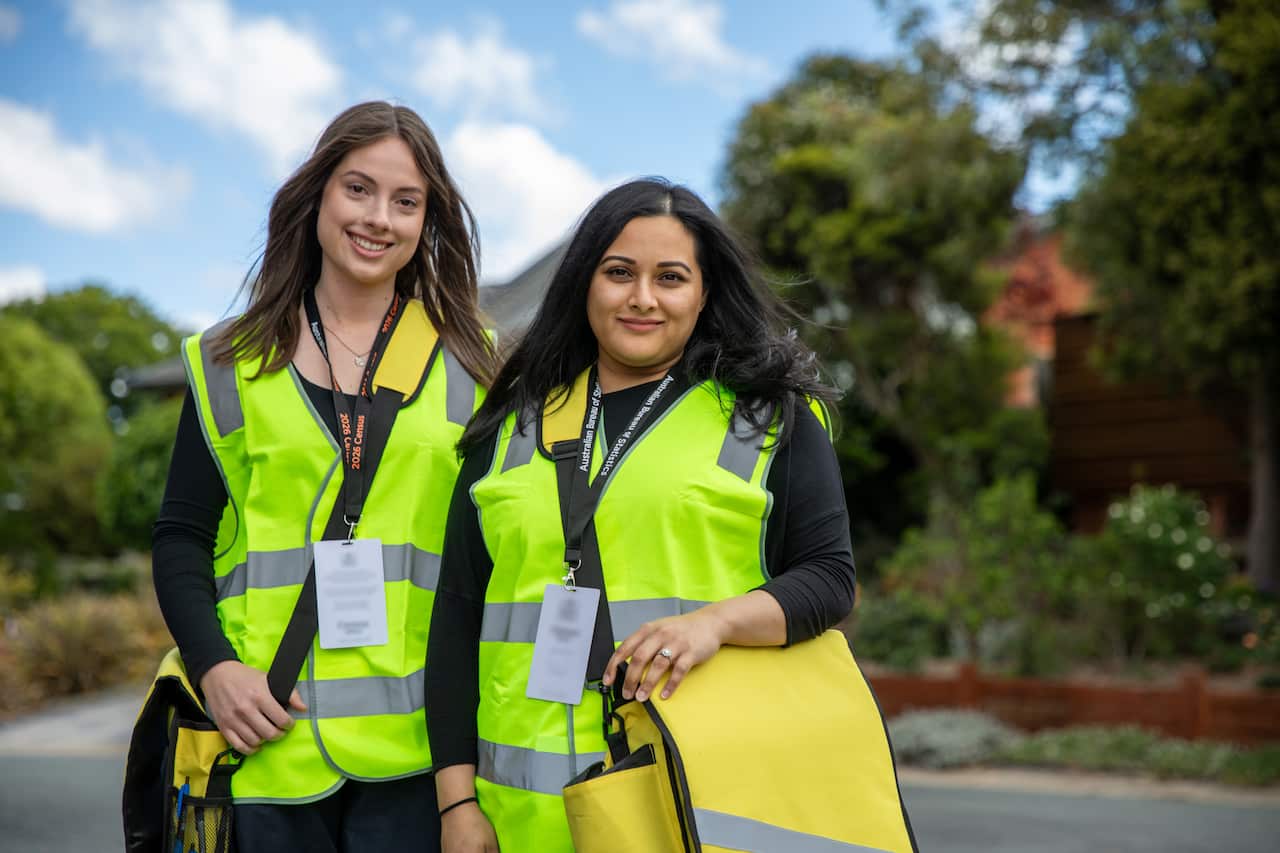 Two women wearing a fluorescent yellow vest, a government lanyard and yellow bags stand smiling.