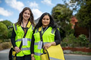 Two women wearing a fluorescent yellow vest, a government lanyard and yellow bags stand smiling.