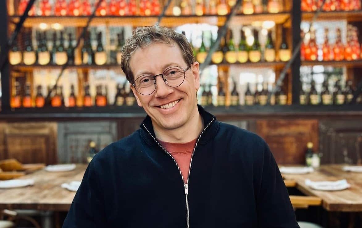 A man with dark, wire-rimmed glasses smiles. You can see bottles of wine and restaurant tables behind him. 