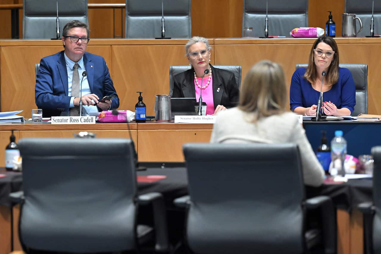 Three senators seated at a desk in front of microphone listen as a woman seated opposite them speaks.