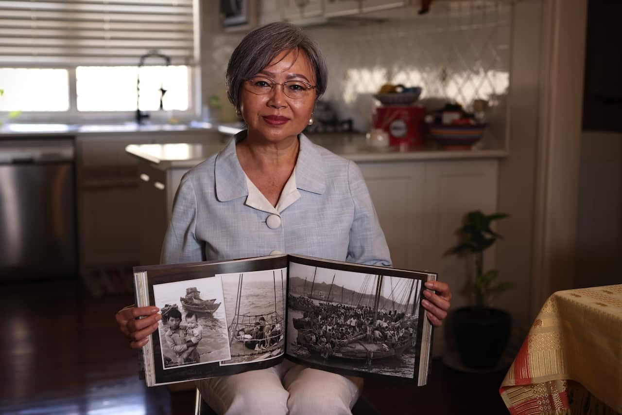 A woman sitting in a kitchen and wearing a light blue shirt and white pants holds an open books with black and white pictures of boat people on each page.