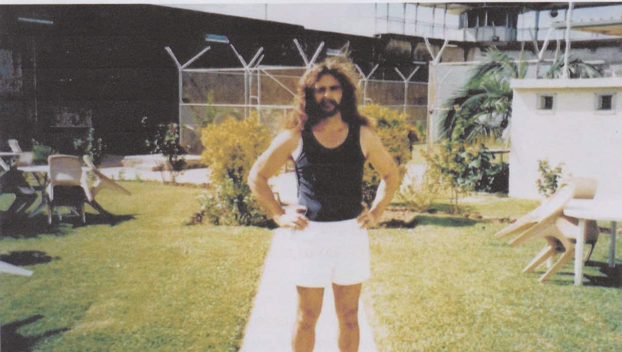 A man with a beard and long hair and wearing a vest and pair of shorts stands in a garden with his hand on his hips.