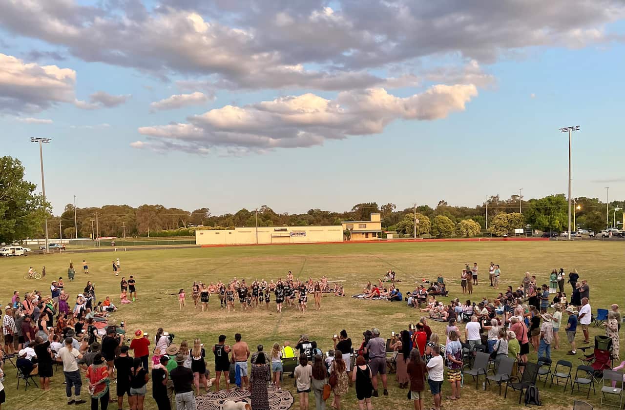 A photo from above showing hundreds of people gathered on an oval, with most people standing and sitting in a large semi-circle around a group of culturally painted Gomeroi dancers.