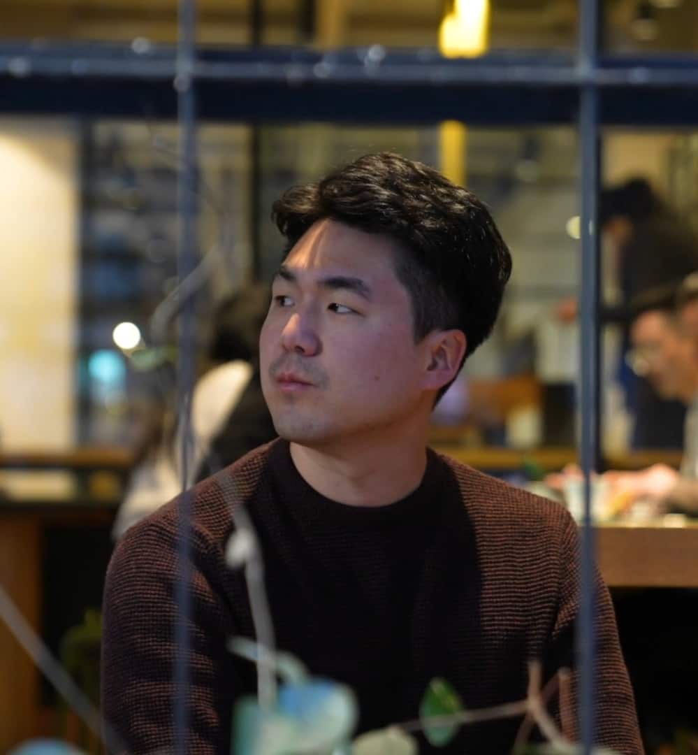 A man in a black t shirt and brown cardigan looking pensively off to his right side