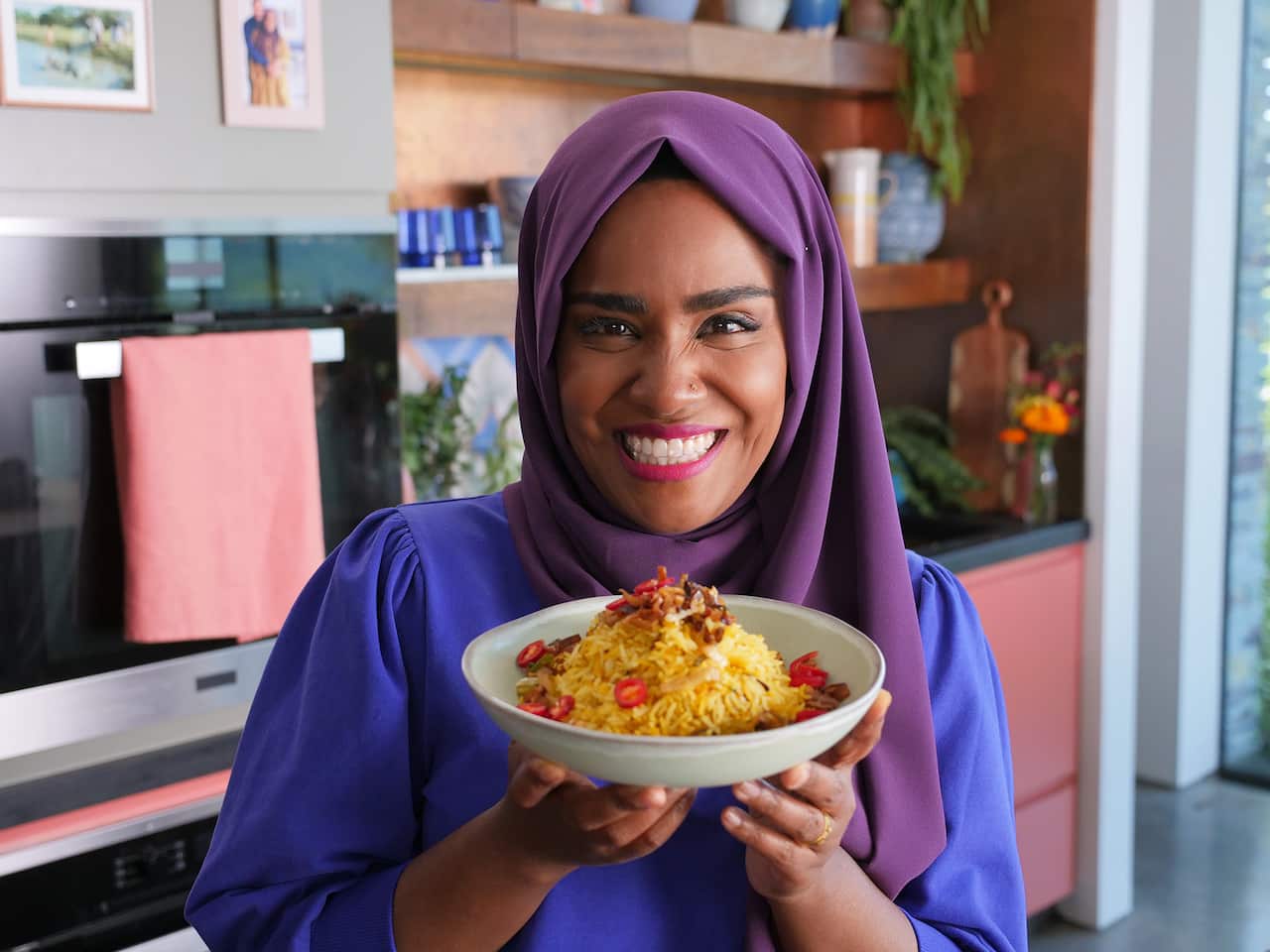 Nadiya stands in her kitchen, smiling, and holding a bowl biryani.
