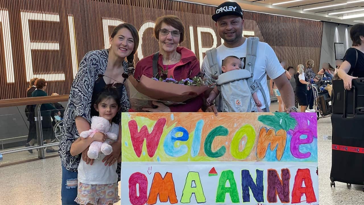 A young couple with two young kids welcomes their grandma at the airport, holding a sign that says 'Welcome Oma Anna'.