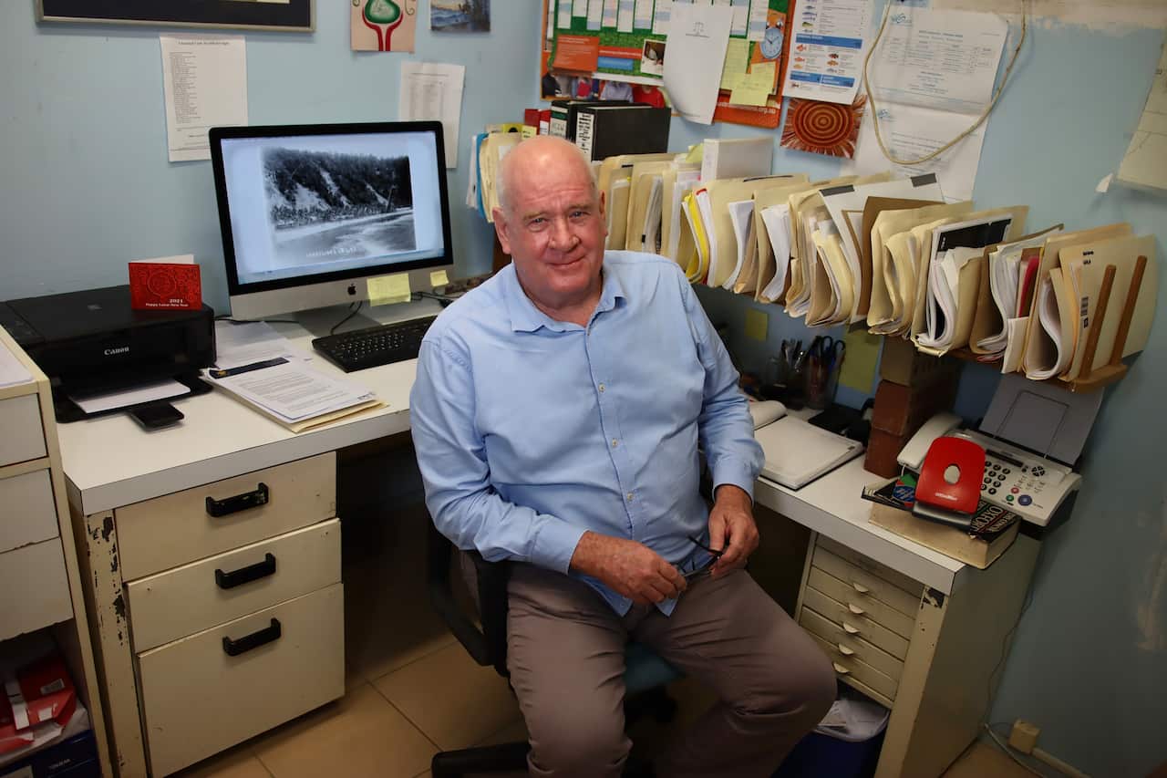 A middle-aged man in a light blue shirt sits in front of a computer in an office.