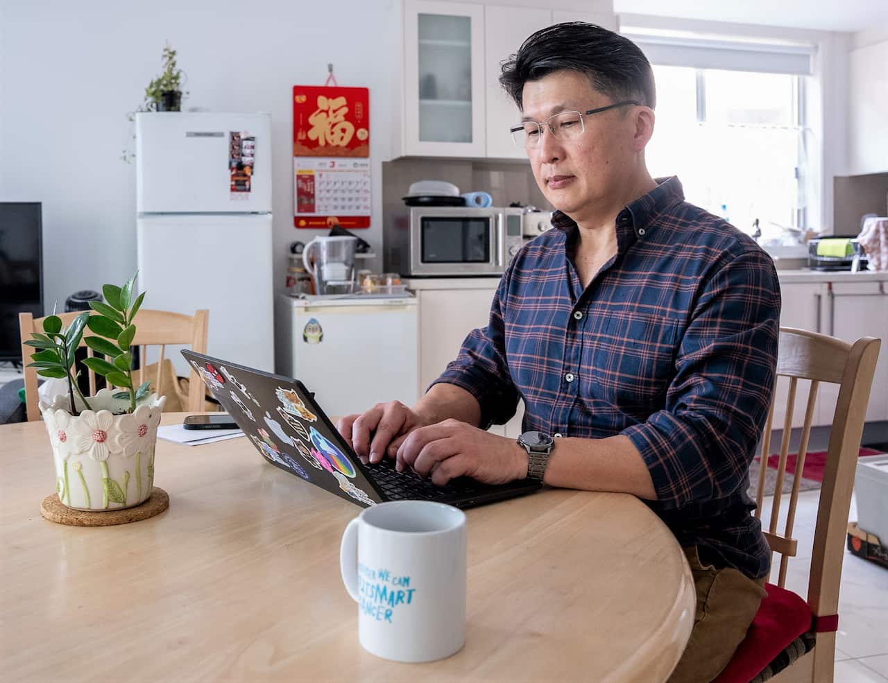 A man in a blue checkered shirt sits at a table using a laptop.