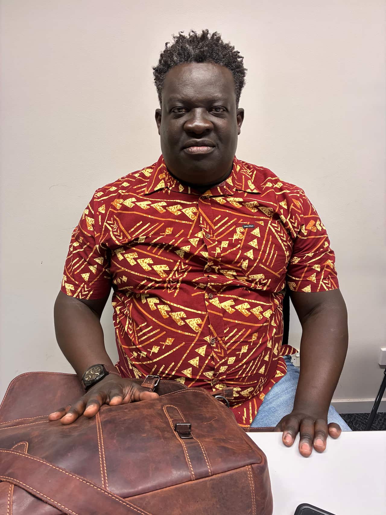 A middle-aged black man wearing a red-and-yellow patterned shirt sits in an office.