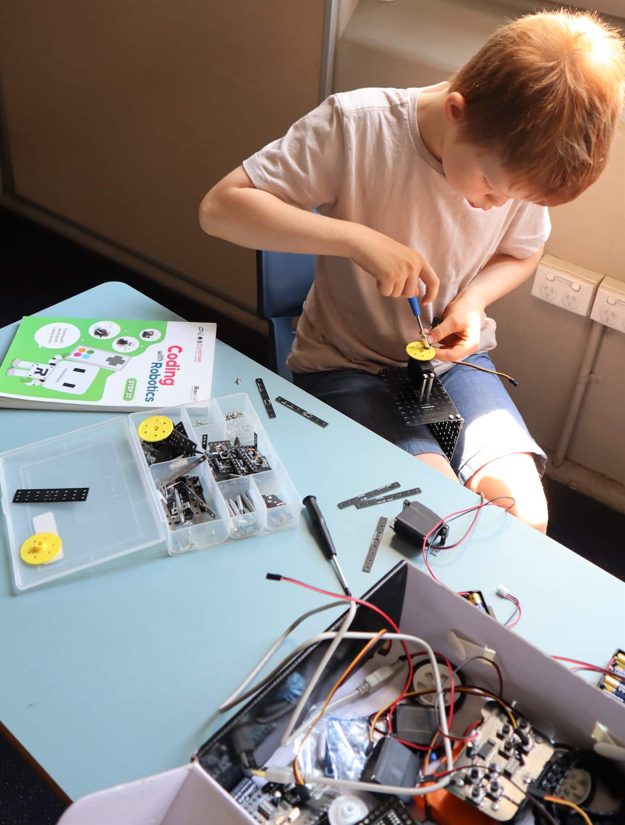 A young boy using a screwdriver on a robot component. He is sitting at a blue desk and there is a box of robot components on it.