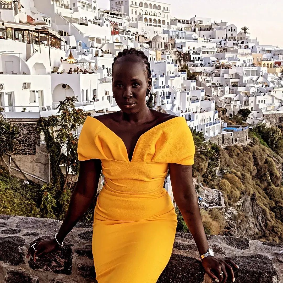 A woman with braided hair and a bright yellow dress leans against a rock, the white buildings of Santorini sprawled behind her.